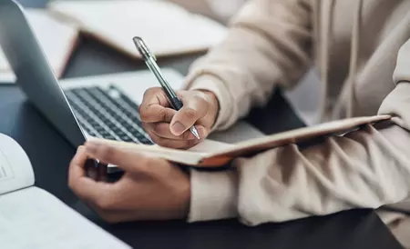 Close-up image of a person's hands taking notes in front of their laptop 